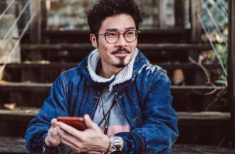 [Featured] A college student in a denim jacket sits on some steps outside while looking at his cellphone.