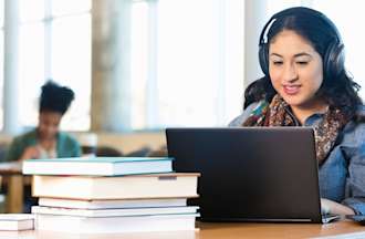 [Featured Image] A machine learning student works on a machine learning project on their laptop in a library at a wooden table. They're wearing headphones and there is a stack of books next to their computer.