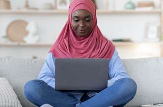 Woman wearing a pink al-amira sits on a grey sofa working on a laptop computer