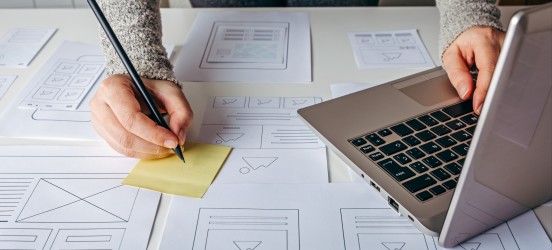 [Featured Image] Overhead view of a hand taking notes next to a laptop computer.