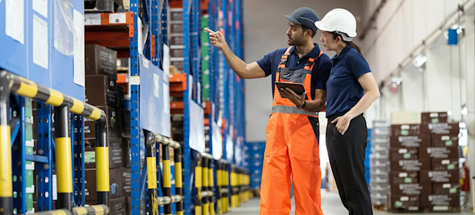 [Featured Image] A warehouse foreman and manager are having a discussion in a warehouse while one is holding a digital device that uses AI in logistics.

