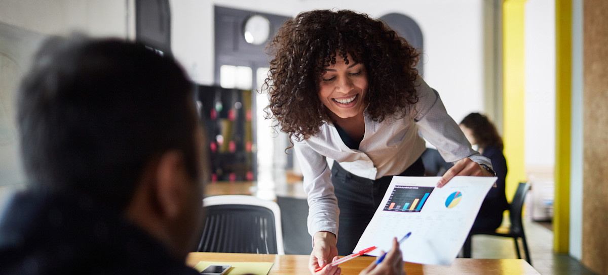 [Featured image] Person in an open office space smiles as they lean over a desk to share their new data findings with their colleague.