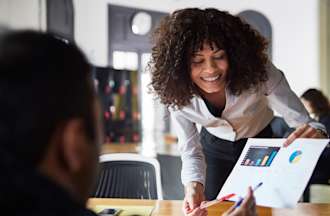 [Featured image] Woman in an open office space smiles as she leans over a desk to share her new data findings with her colleague.