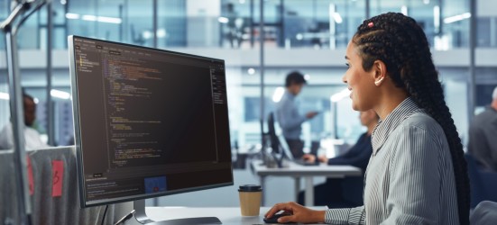 [Featured Image] An AI engineer with long dark hair in a gray striped shirt works in front of their computer screen in a shared office with lots of windows.  