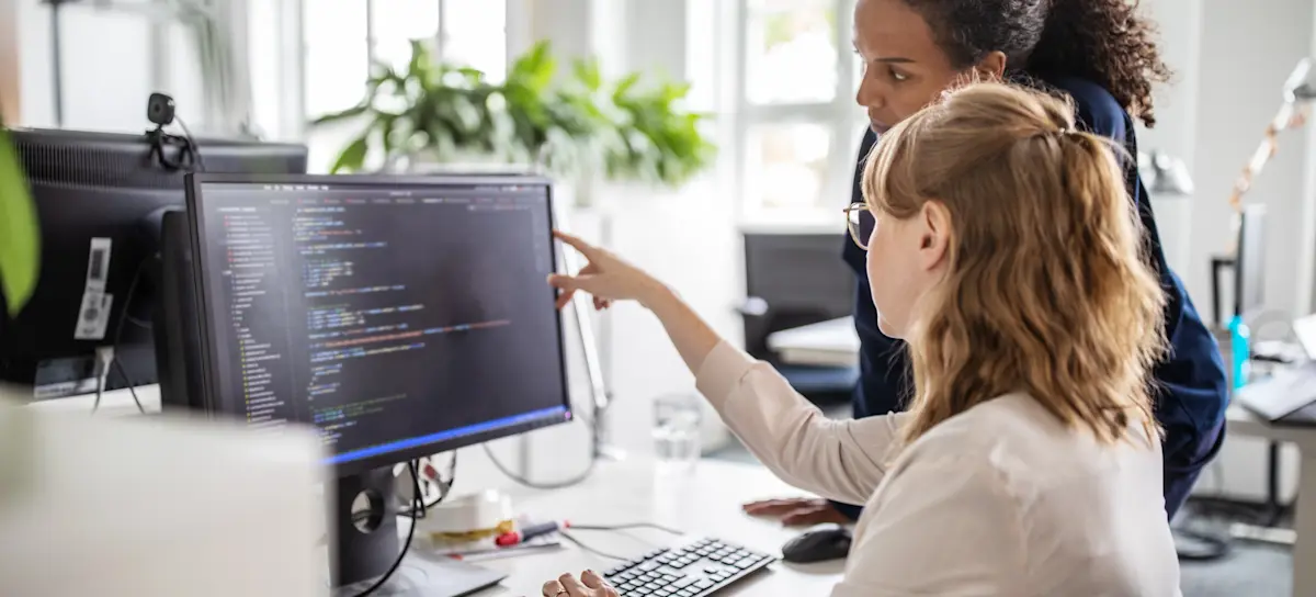 [Featured Image] Coworkers discussing the concept of serialization in an office environment, one explaining the process while pointing at a computer screen displaying code.
