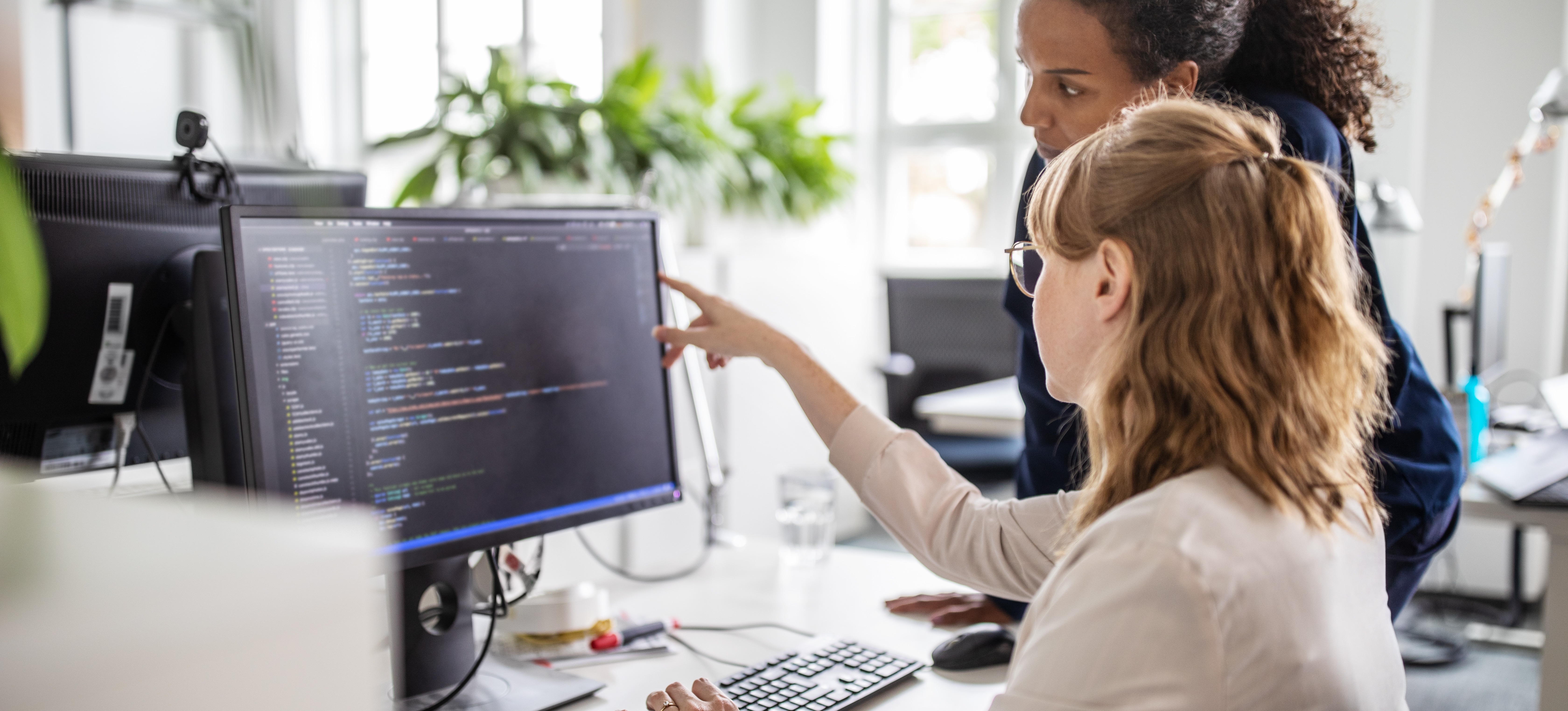 [Featured Image] Coworkers discussing the concept of serialization in an office environment, one explaining the process while pointing at a computer screen displaying code.
