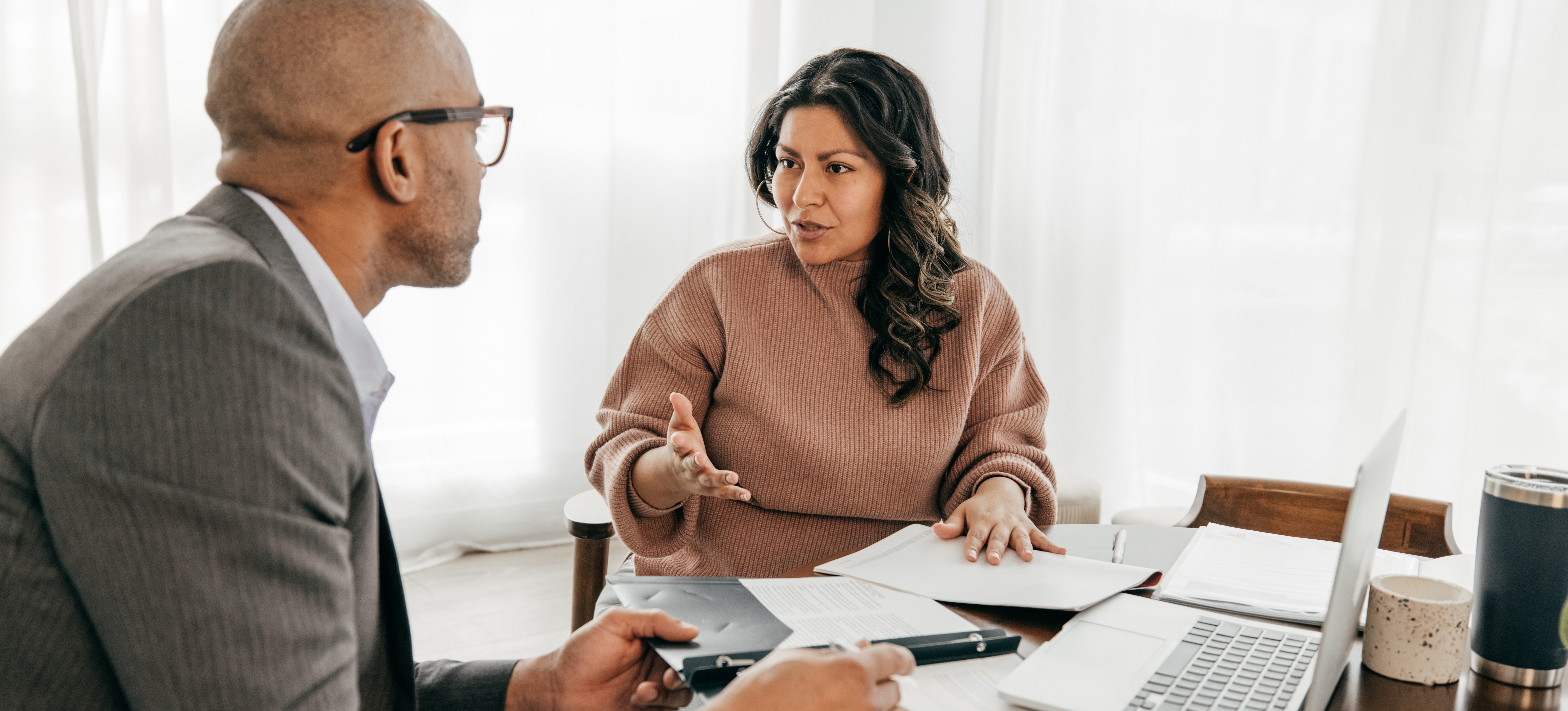 [Featured Image] A business consultant in a brown sweater explains what their role is to a potential new client sitting at their desk in front of an open laptop.

