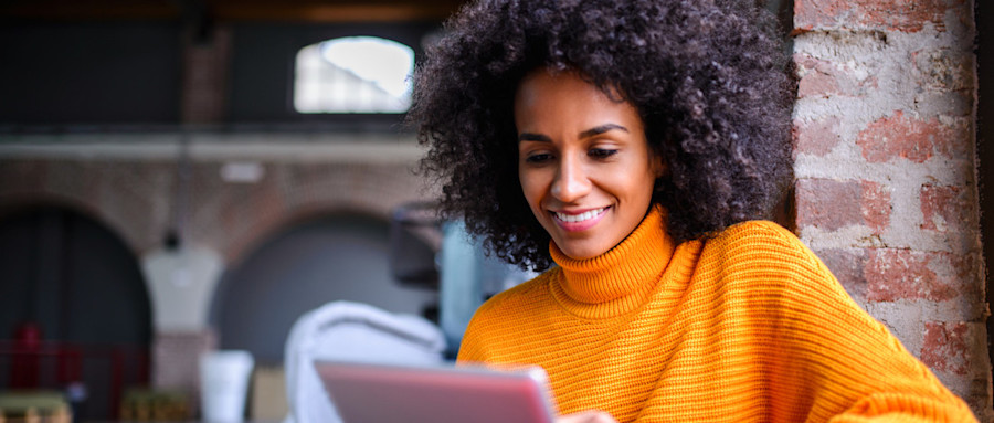 [Featured Image] An Executive MBA candidate wearing a turtleneck sweater in a large brick room smiles while looking down at their tablet.
