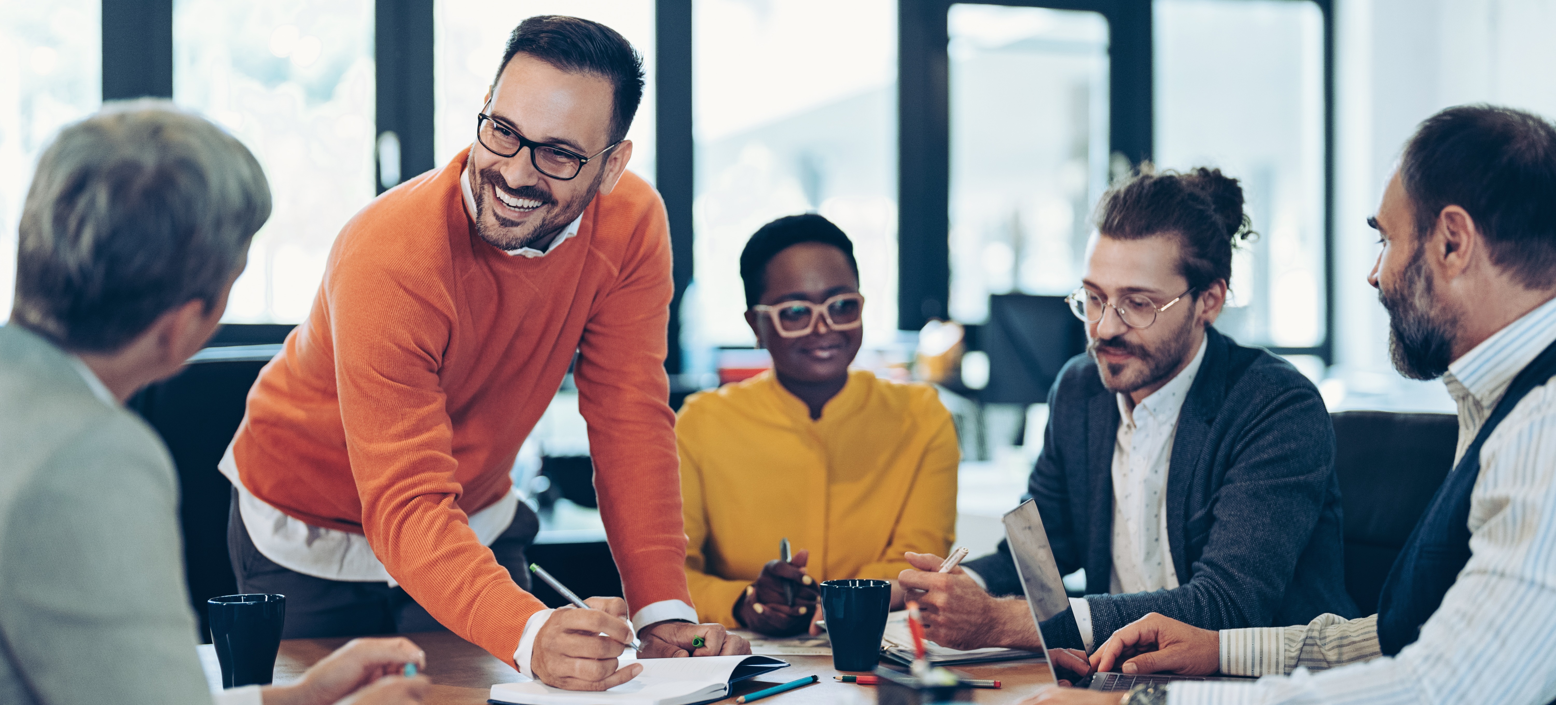 [Featured image] A smiling, positive programme manager stands at a work table with a pen in his hand and explains a concept to a team member as the rest of the team discusses items while sitting at the table in an office setting.
