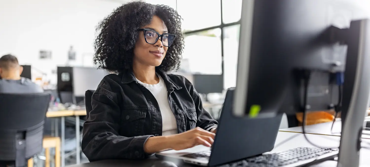 [Featured image]: A person sits at a desk working on a laptop and desktop computer after earning a DevOps foundation certification.
