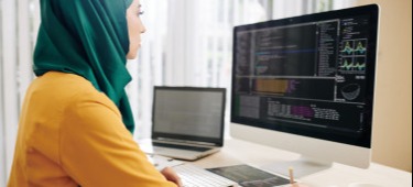 [Featured Image] Female data engineer sits in front of a dual computer screen, looking at data visualizations and writing in a notebook.