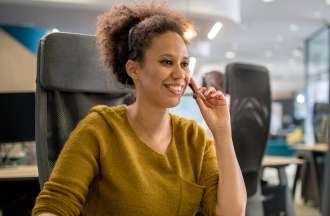 [Featured Image] A customer service representative answers customer questions over the phone. They are seated at a desk facing a computer monitor and wearing a telephone headset.