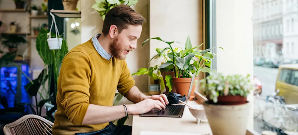 [Featured Image] A freelancer works in a coffee shop, researching new freelance opportunities on their laptop. 