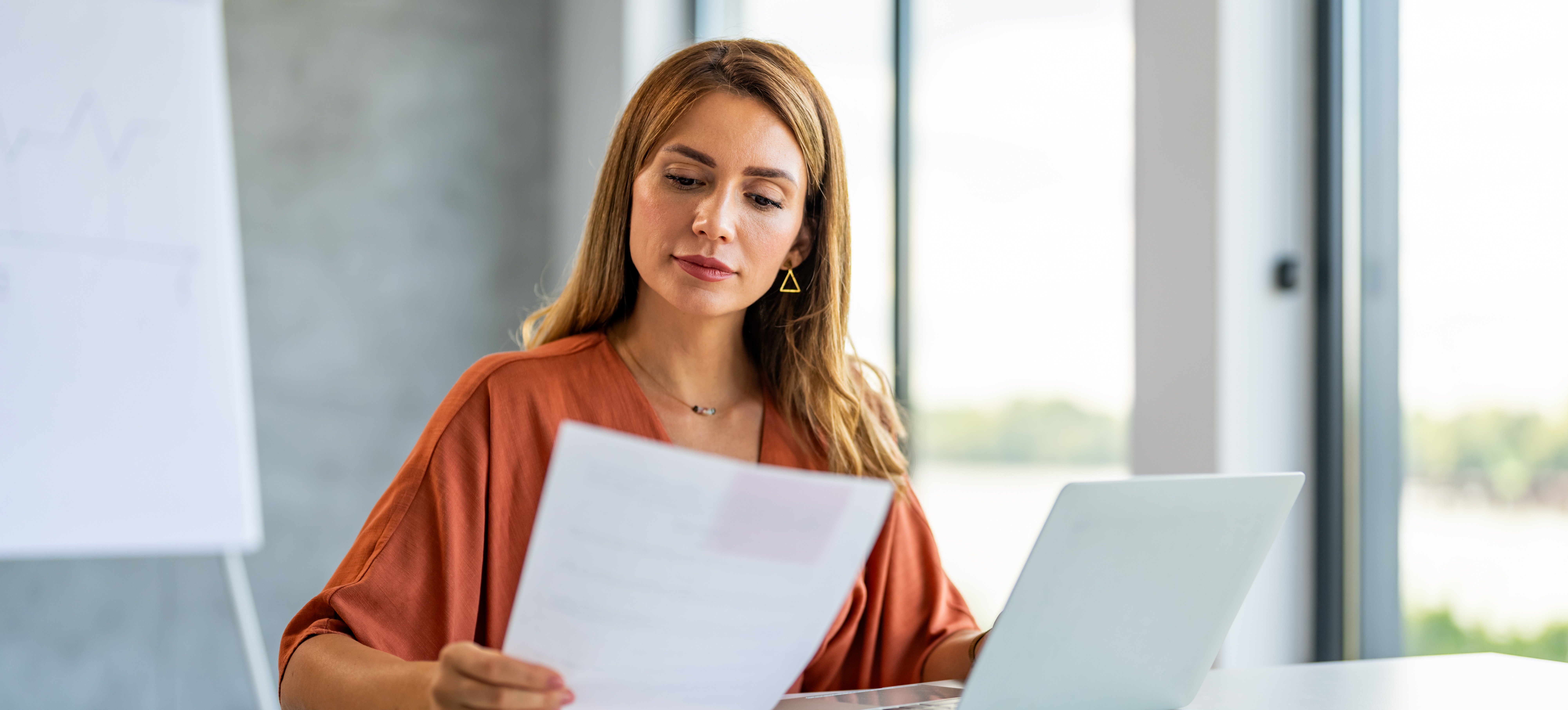 [Featured Image] A person holds their cover letter in their hand and reads it while sitting in front of their laptop and contemplating if cover letters are necessary.
