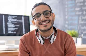 [Featured image] A young man in glasses and a burnt orange sweater sits in front of his desktop computer. There's a chalkboard in the background with lots of writing on it. 