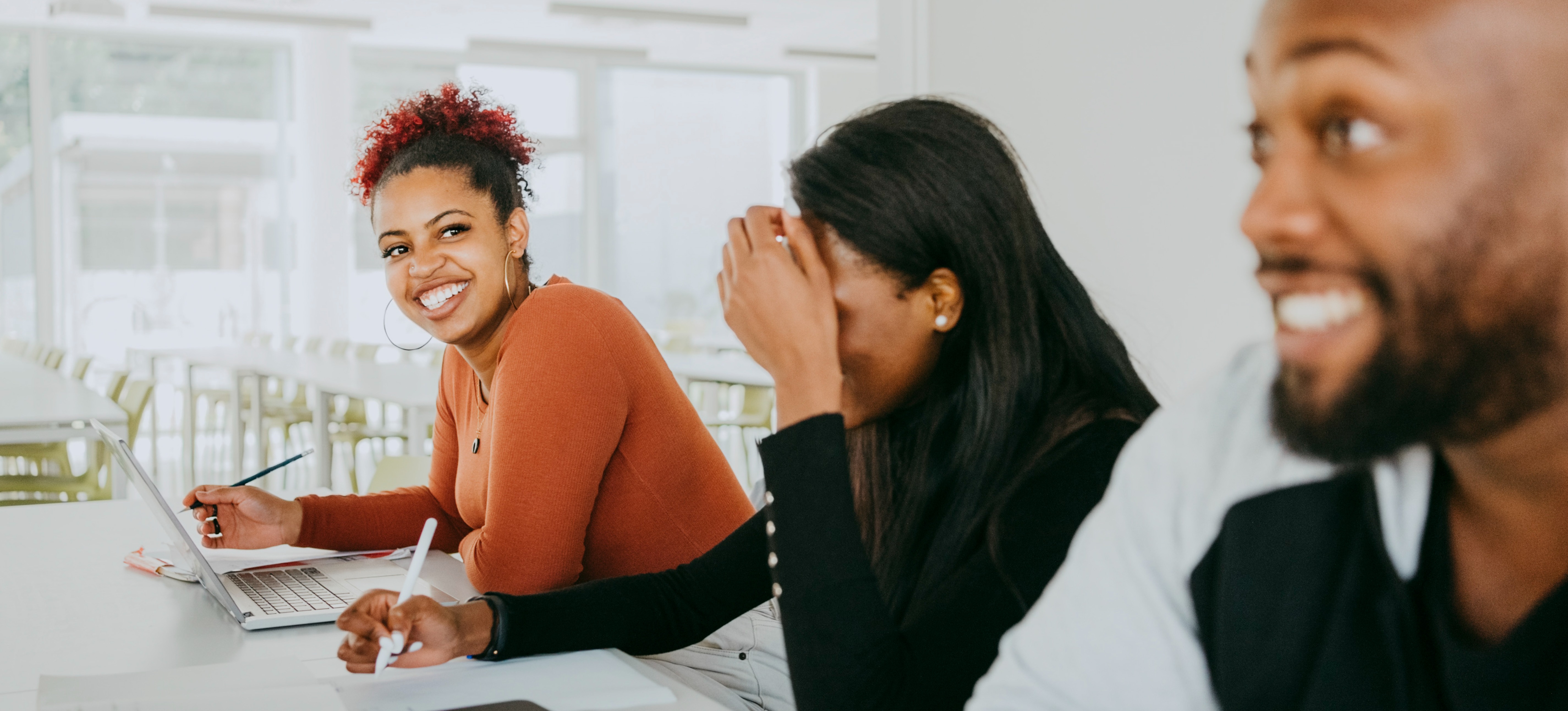 [Featured Image] Three smiling multiracial professionals sitting at a table taking notes on paper and laptops.