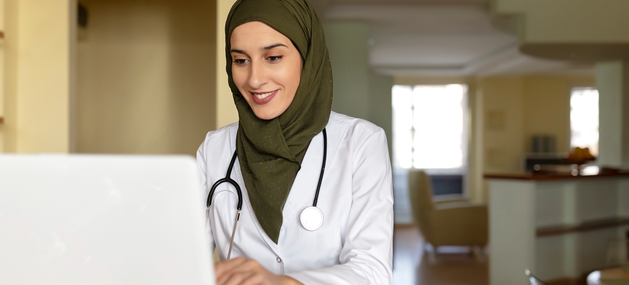 {Featured Image] A person wearing a hijab, a white lab coat, and a stethoscope sits using a laptop.  