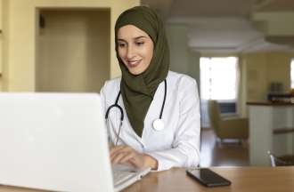 A woman wearing a hijab, a white lab coat, and a stethoscope sits using a laptop.  