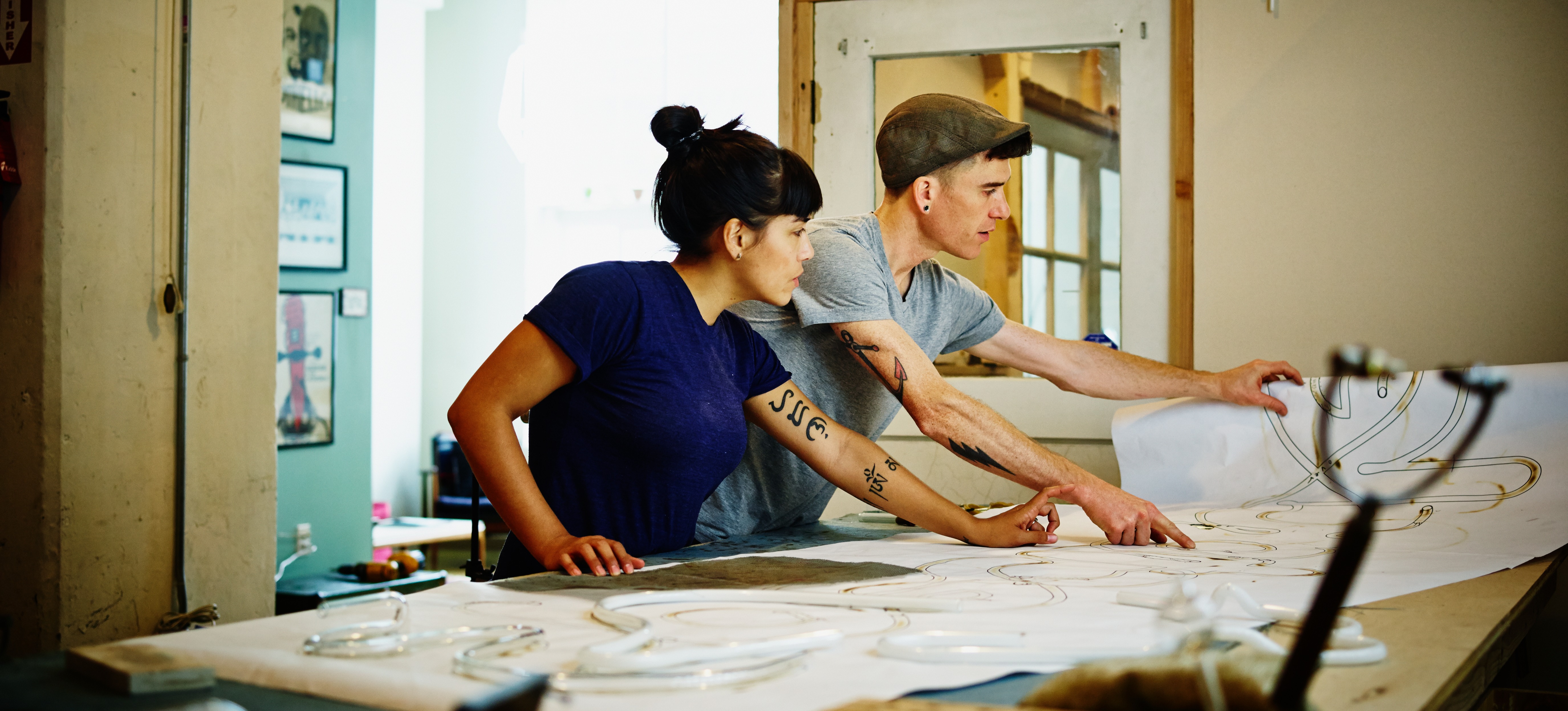 [Featured Image] Two creative designers stand at a large work table and handle large pieces of paper with hand-drawn designs.
