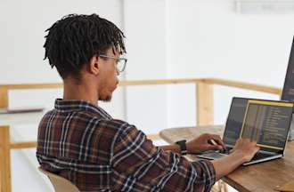 [Featured image] A person wearing a plaid shirt sits at a desk and reviews code using a laptop and a separate display monitor.