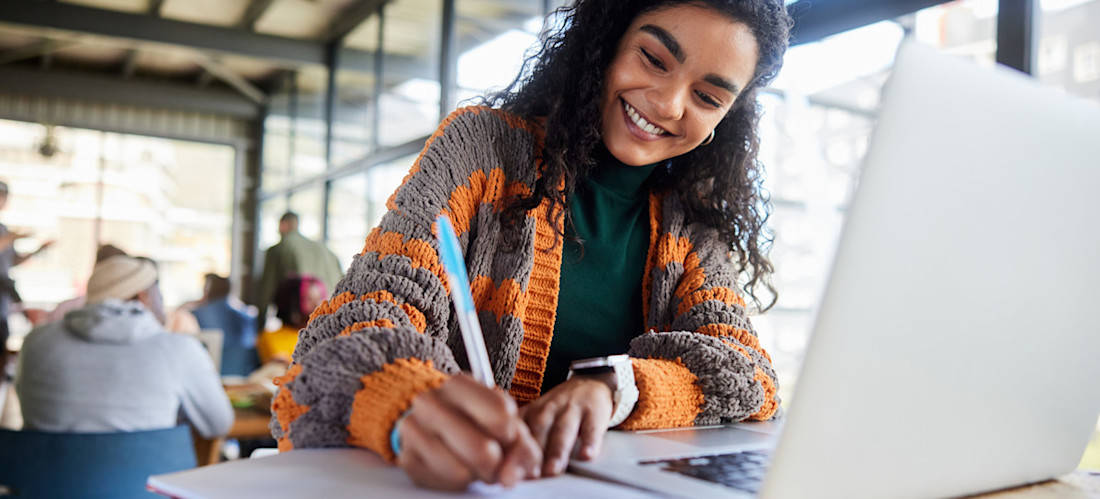 [Featured Image] A young college student writing notes and working on a laptop while doing their homework in a busy campus cafeteria.
