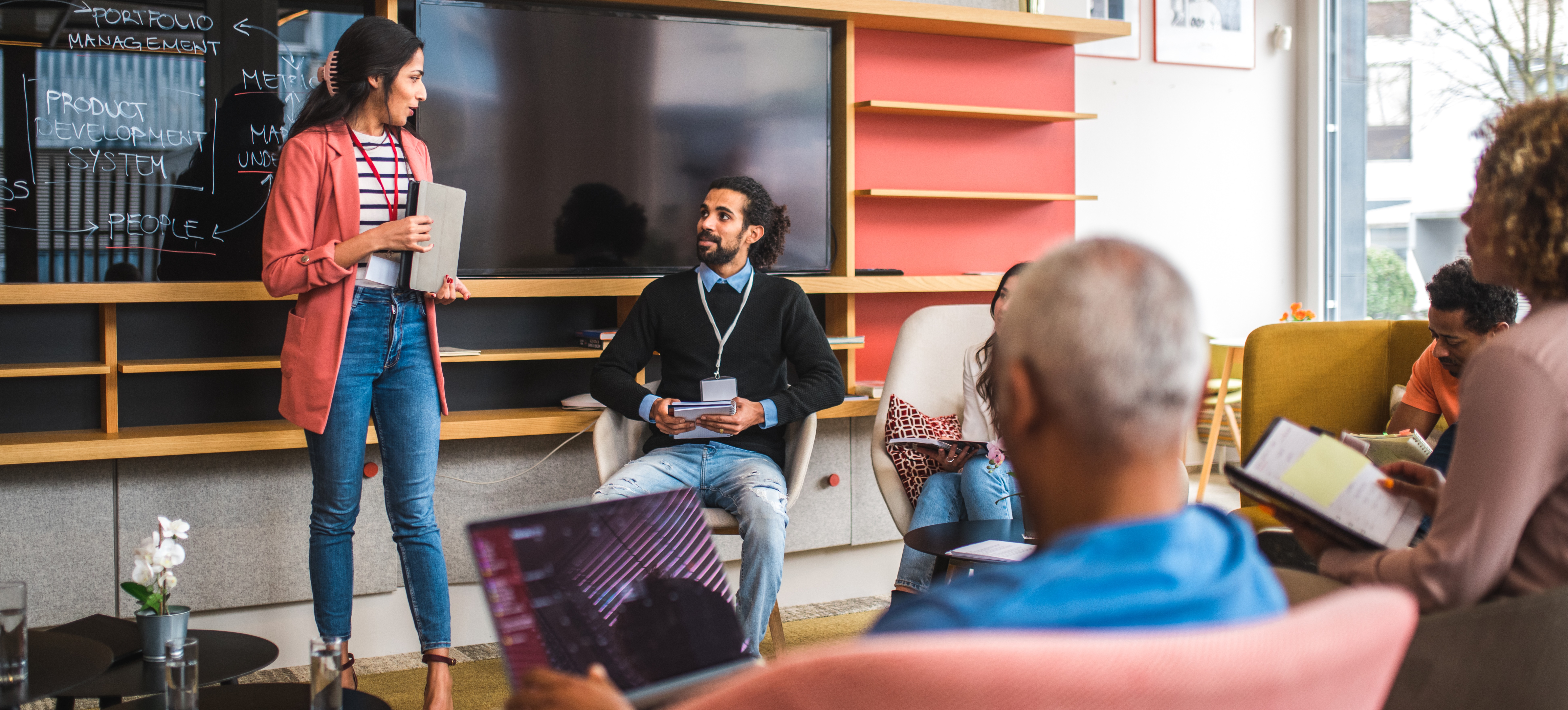 [Featured Image] A sales operations manager addresses the sales team gathered around her in a casual and colorful office setting.
