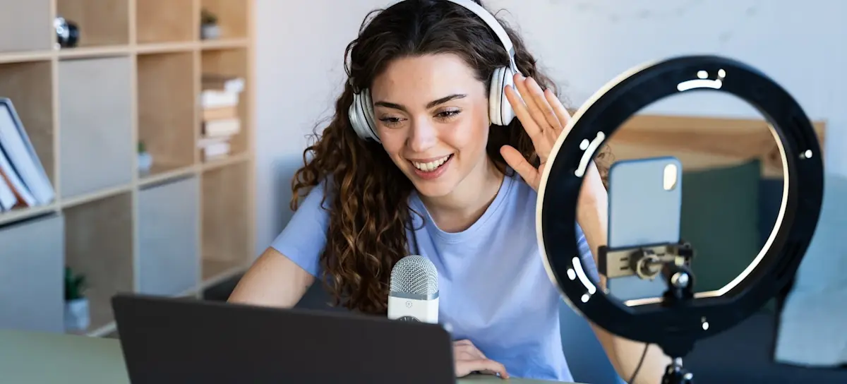 [Featured Image] After providing an impressive digital content creator resume, a newly-hired digital content creator interacts remotely with their team on a laptop with lighting and recording equipment nearby. 
