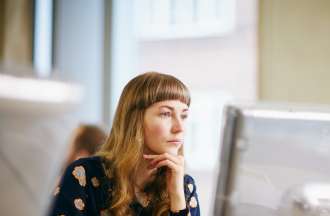 [Featured Image]:  A female, with blonde hair, and wearing a dark patterned dress, is sitting in front of her desktop, in her office. She is sitting by a window. 