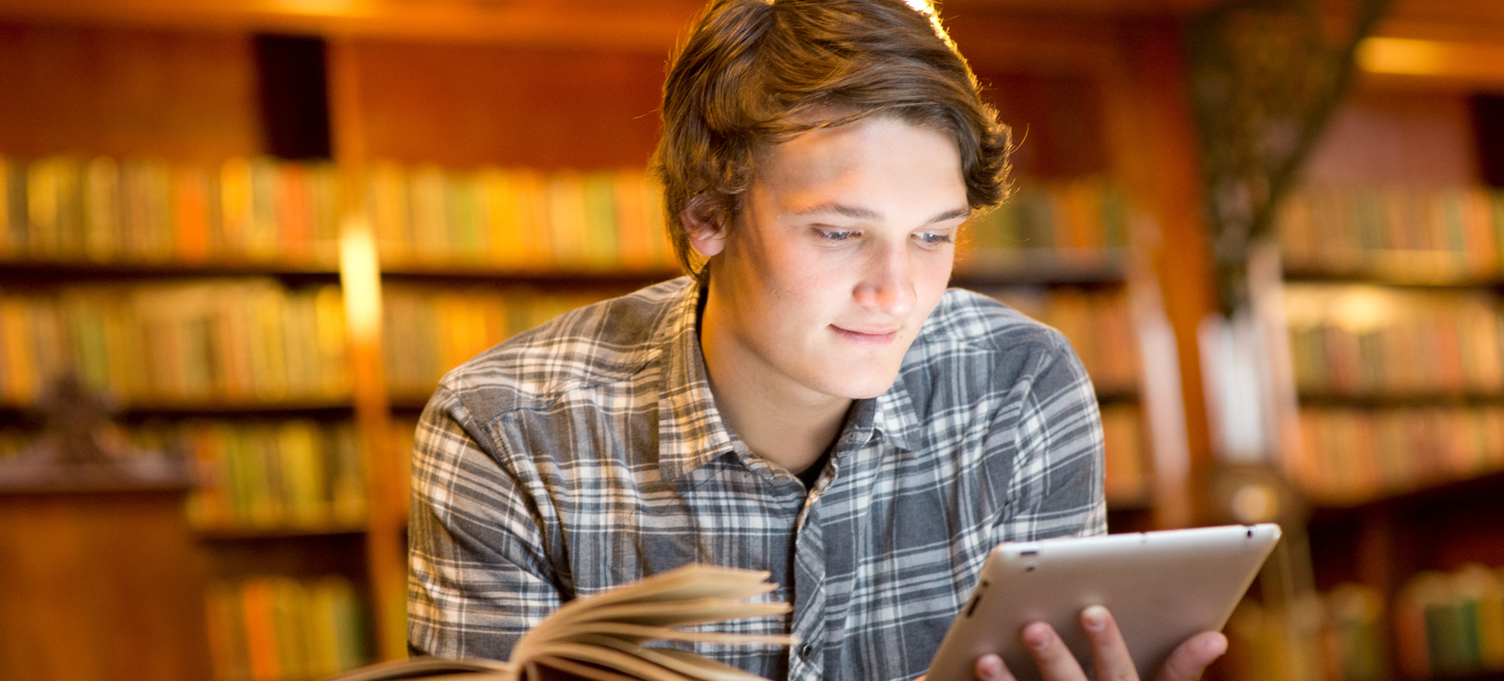 [Featured Image] A learner in a plaid shirt sits in a library at a table with a stack of books, researching adaptive learning platforms on a tablet as they seek the option that best meets their needs. 
