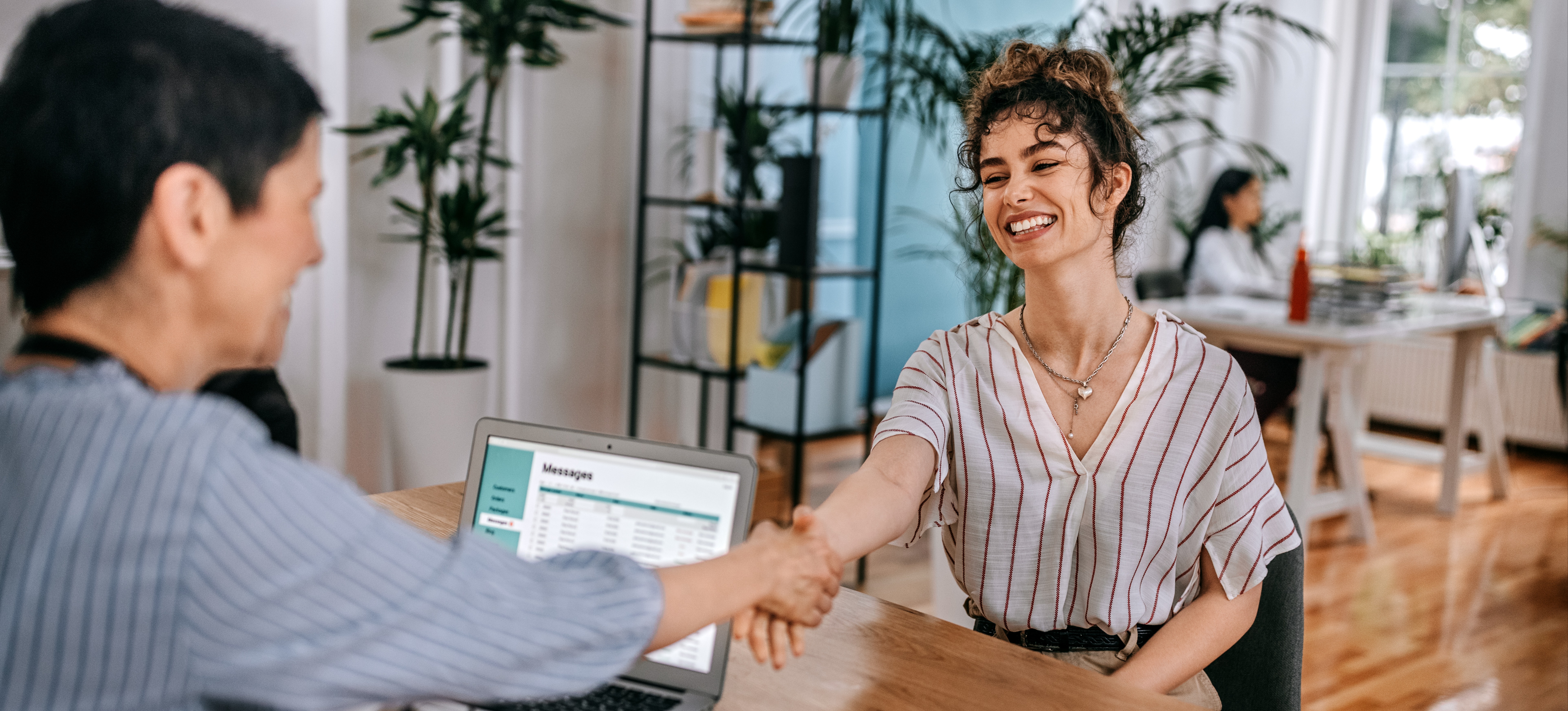 [Featured Image] A young woman learning how to become an HR specialist shakes the hand of another woman over a desk during a job interview.
