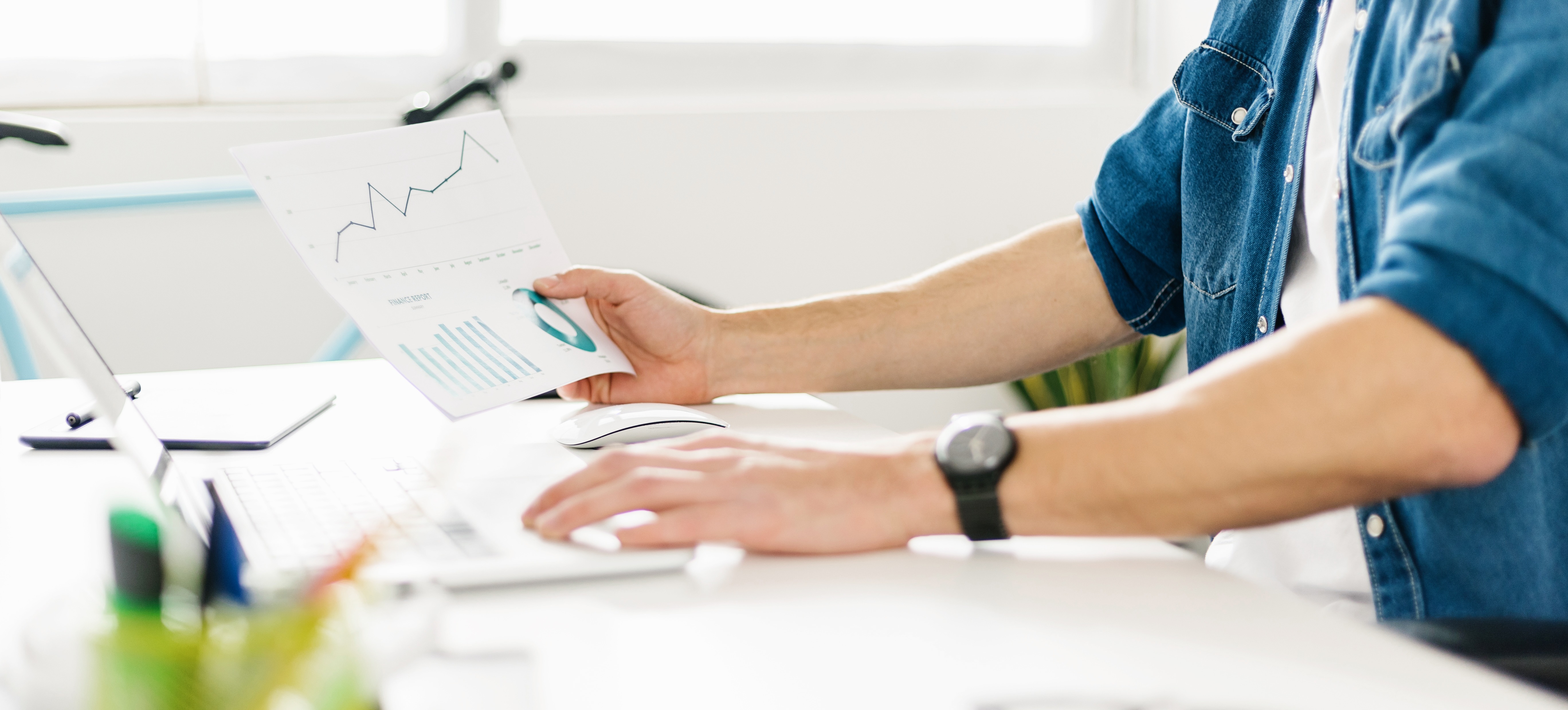 [Featured Image] A digital marketing analyst studies data and graphs at her office desk using printed documents and her laptop.
