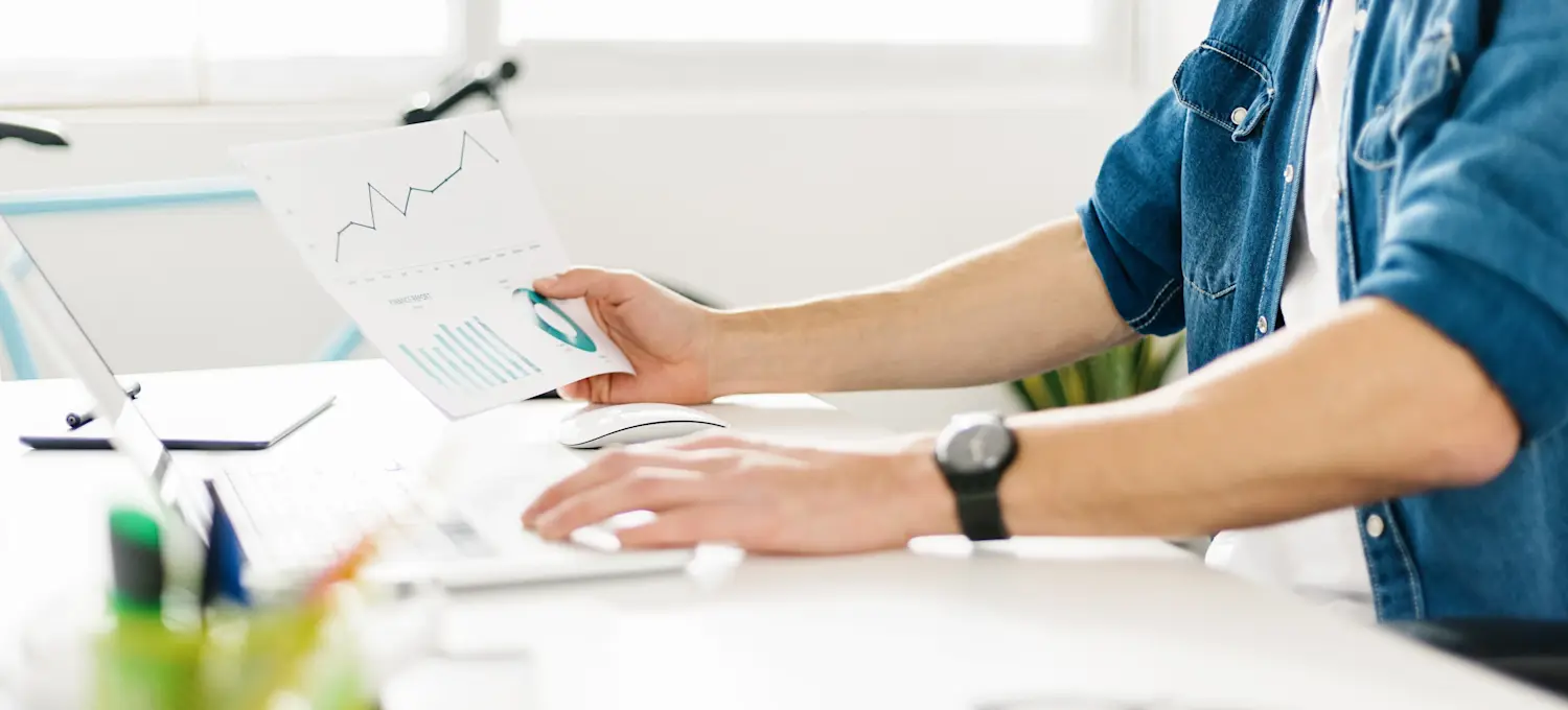 [Featured Image] A digital marketing analyst studies data and graphs at their office desk using printed documents and their laptop.
