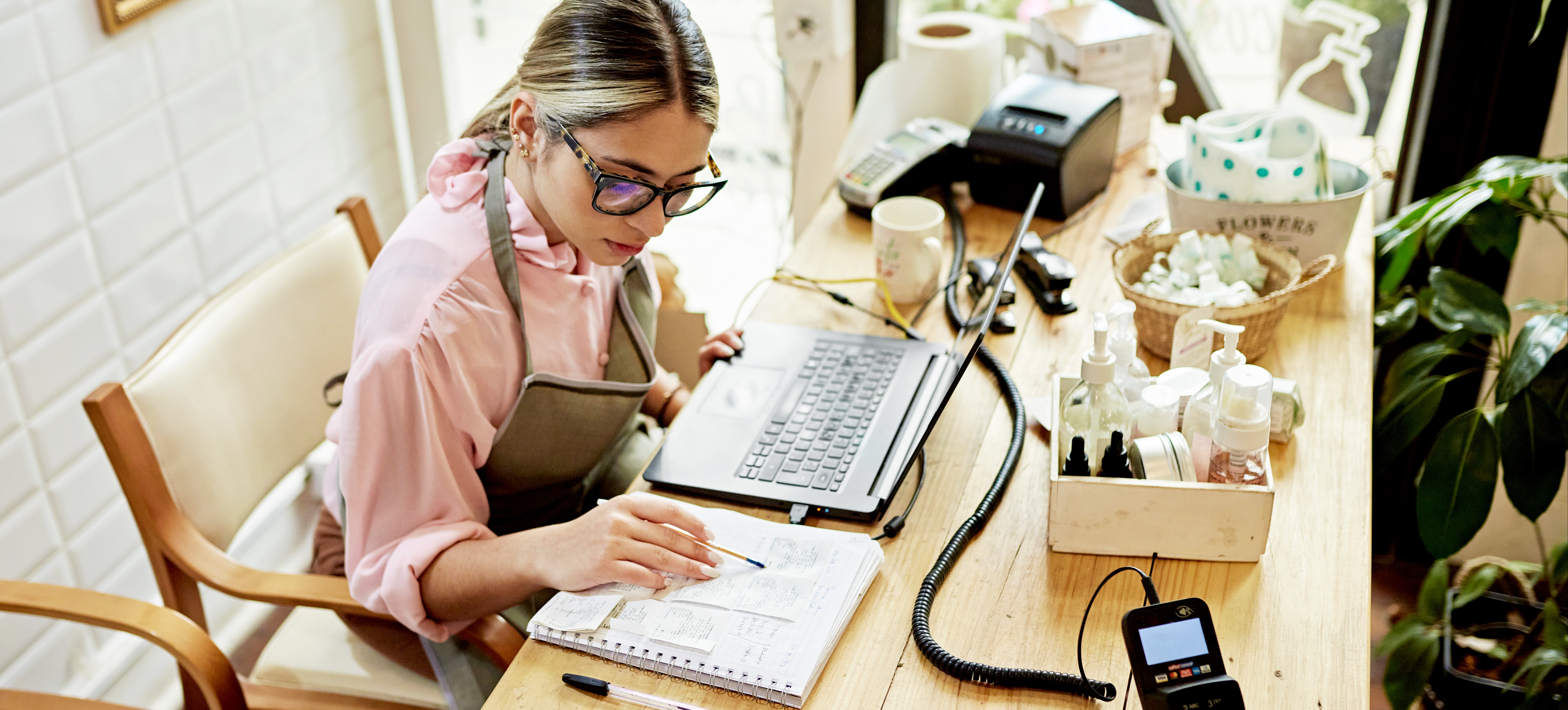 [Featured Image] A young woman sits at her desk with a laptop and notebook, learning small business bookkeeping for her business.