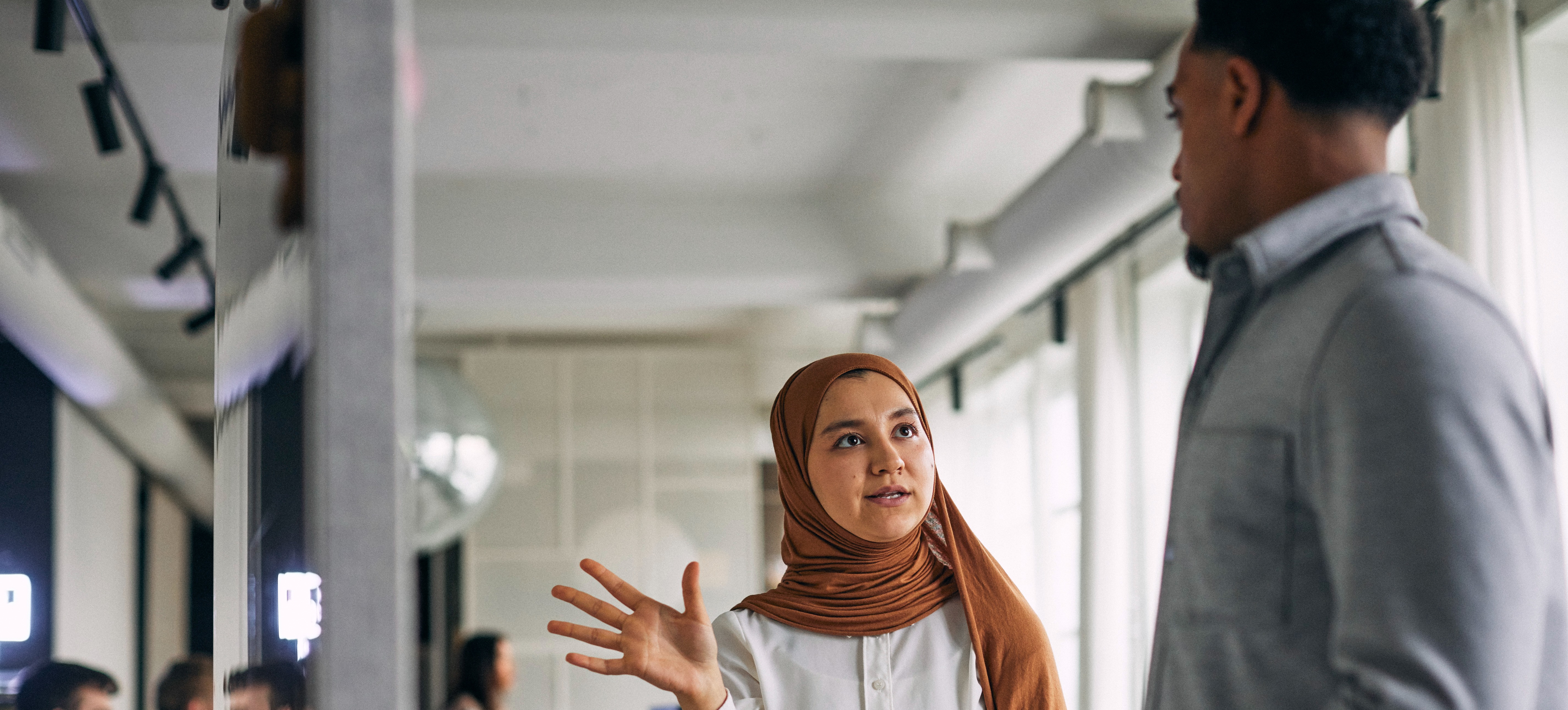 [Featured Image] A businessperson assisting a colleague in front of a computer monitor, while other colleagues work in the background.
