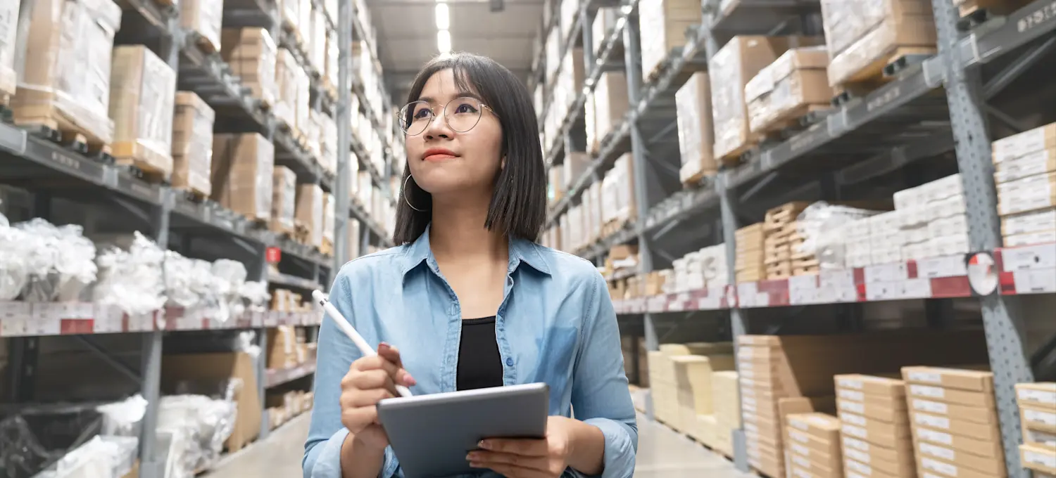 [Featured Image] A person holding a tablet and pen looks up at warehouse shelves full of boxes as they work as a merchandising director, one of the highest-paying retail jobs.
