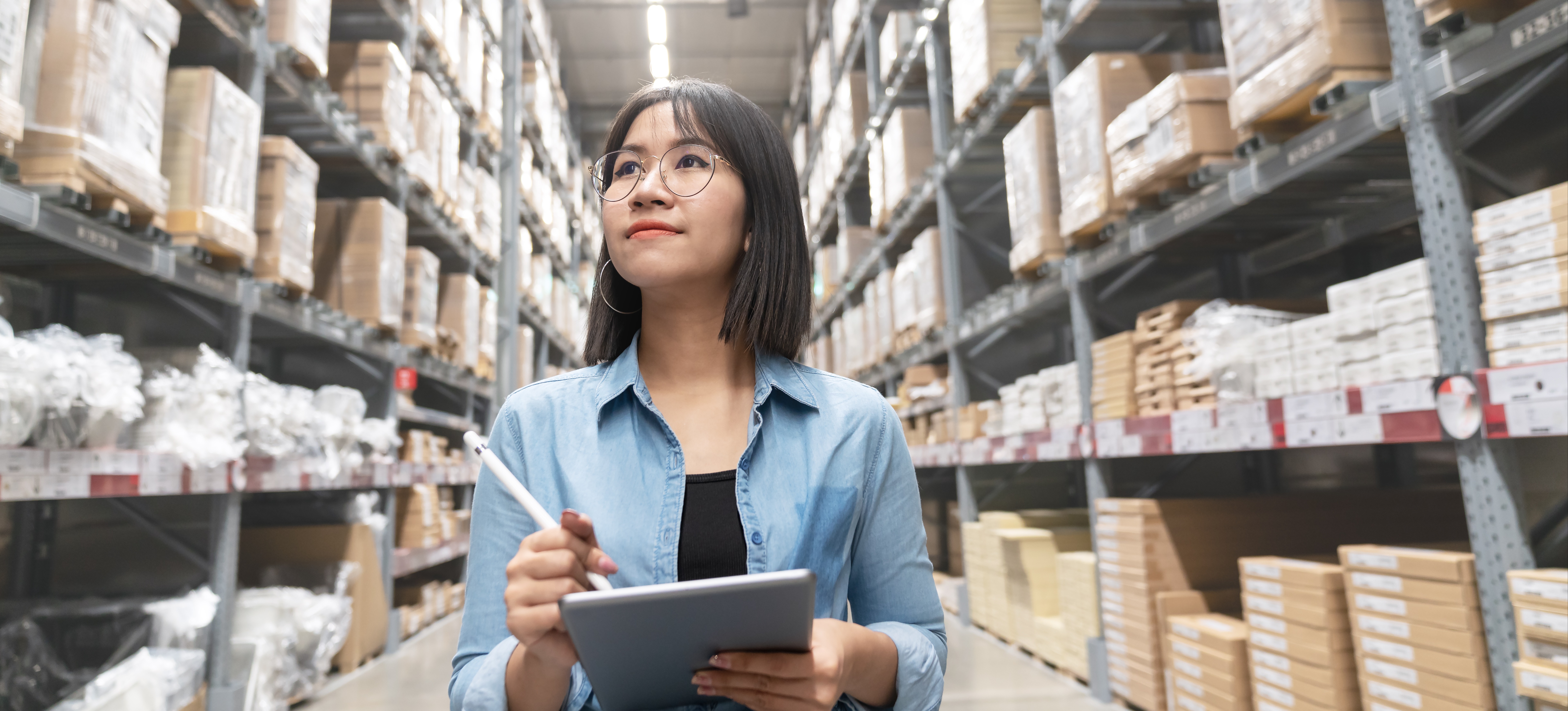 [Featured Image] A woman holding a tablet and pen looks up at warehouse shelves full of boxes as she works as a merchandising director, one of the highest-paying retail jobs.
