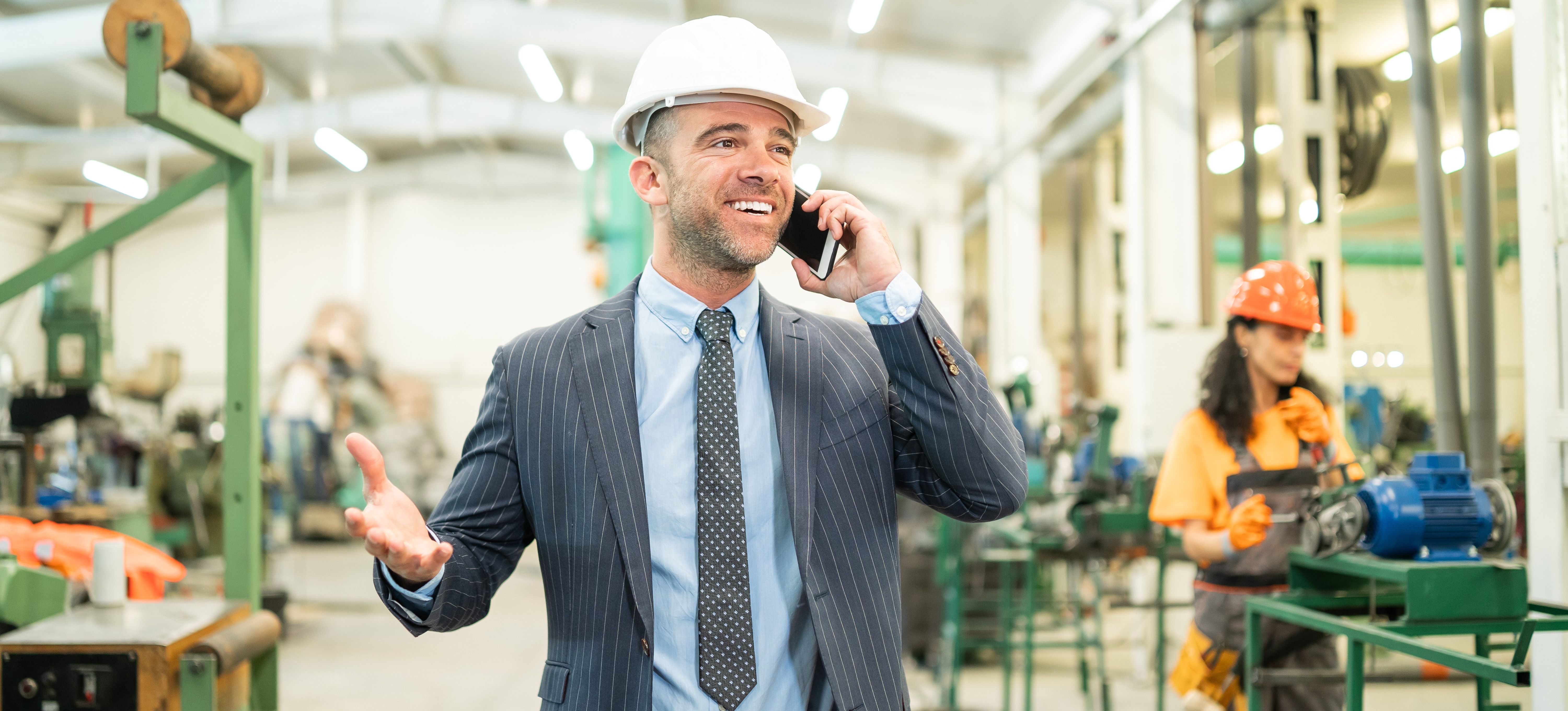 [Hauptbild] Ein Mann, der einen Schutzhelm trägt und für den Lohn eines Vertriebsingenieurs arbeitet, telefoniert während eines Verkaufsgesprächs in einer Fabrik mit einem potenziellen Kunden.
