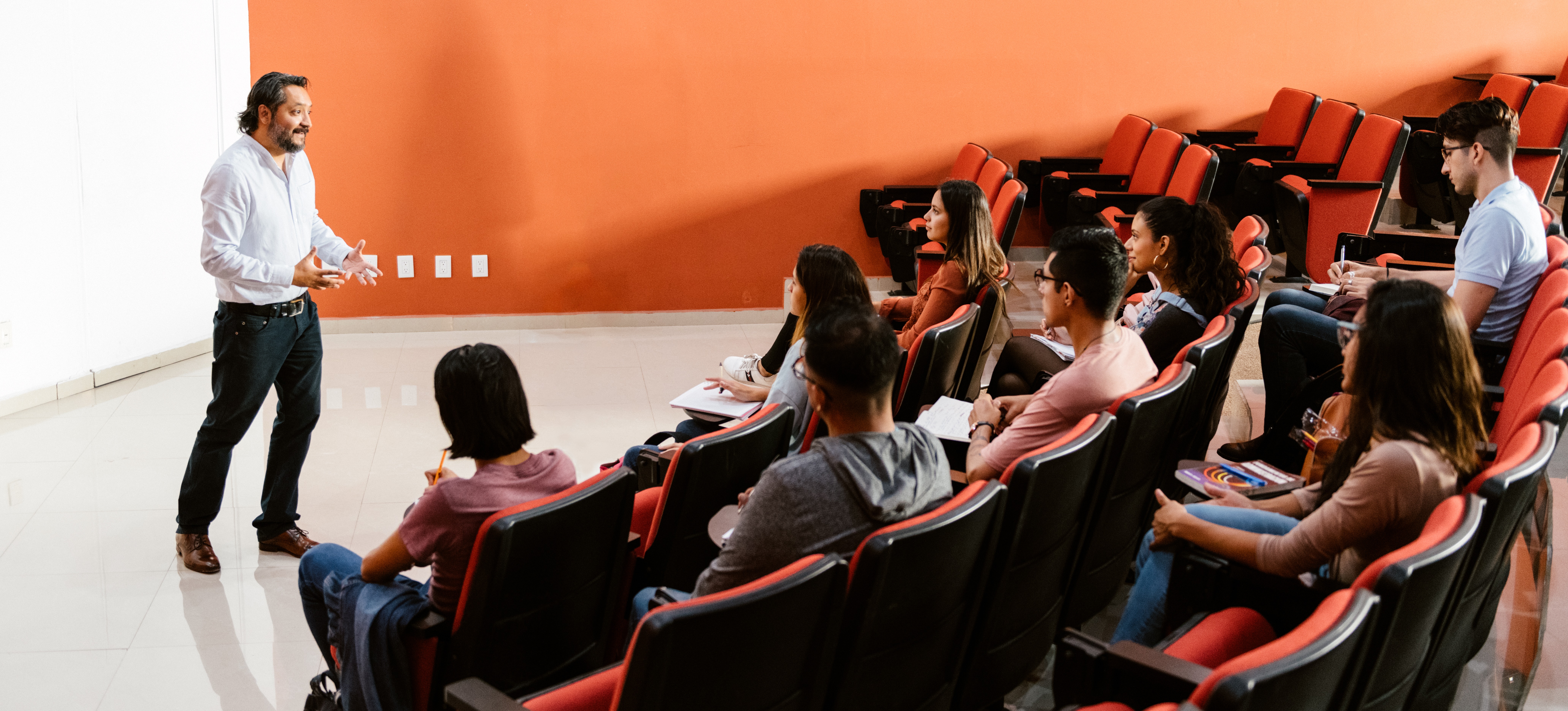 [Featured Image] A male teacher stands infront of a college class teaching after a student asked, "What does gcse stand for?".