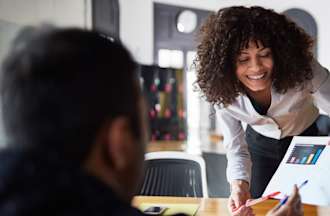 [Featured image] A person in a white blouse leans across a table to show a colleague a printout with data visualizations in order to explain what data science is. 