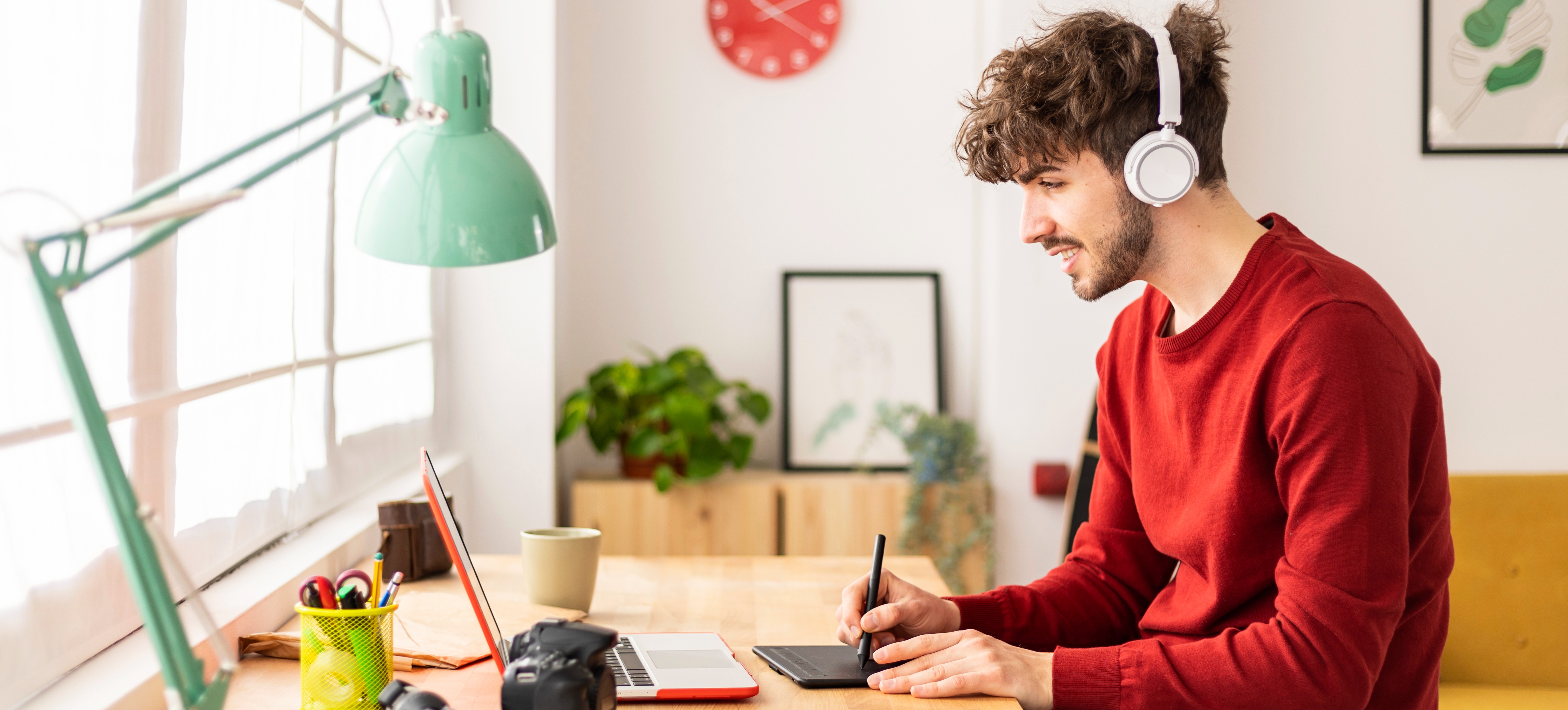 [Featured Image] A graphic designer wears headphones and sits at a desk, using a laptop and tablet to explore new graphic design apps. 
