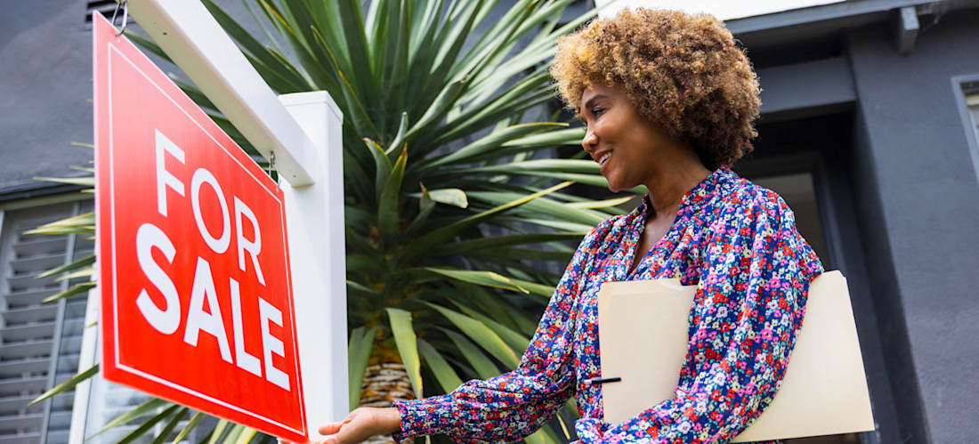 [Featured Image] A real estate agent looks at a for-sale sign outside a property while contemplating how much REALTORS® make.