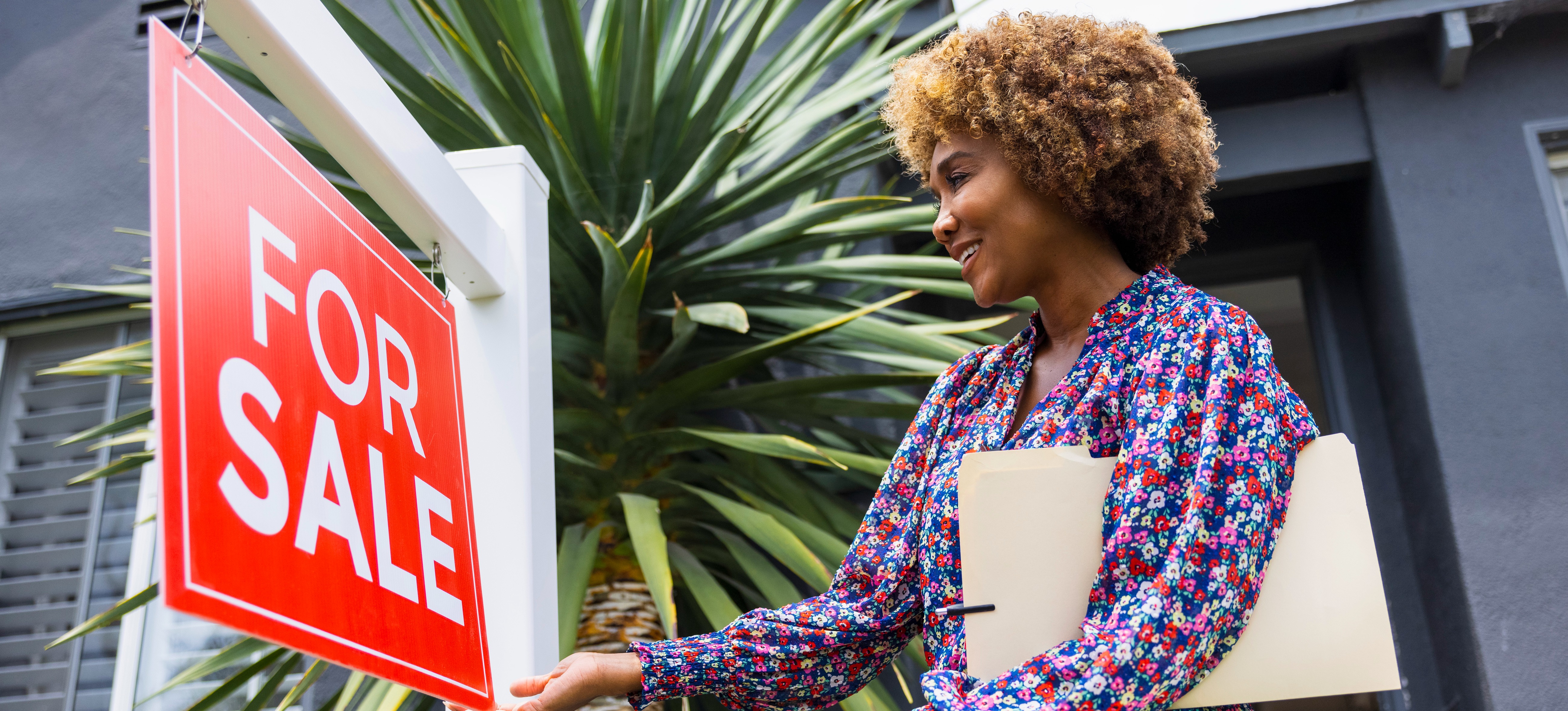 [Featured Image] A real estate agent looks at a for-sale sign outside a property while contemplating how much REALTORS® make.