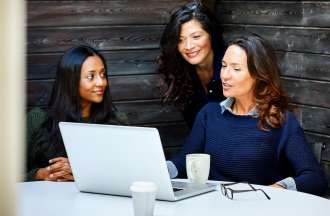 A marketing manager meets with her team at a conference table. One woman has a laptop, glasses, and a coffee cup in front of her.