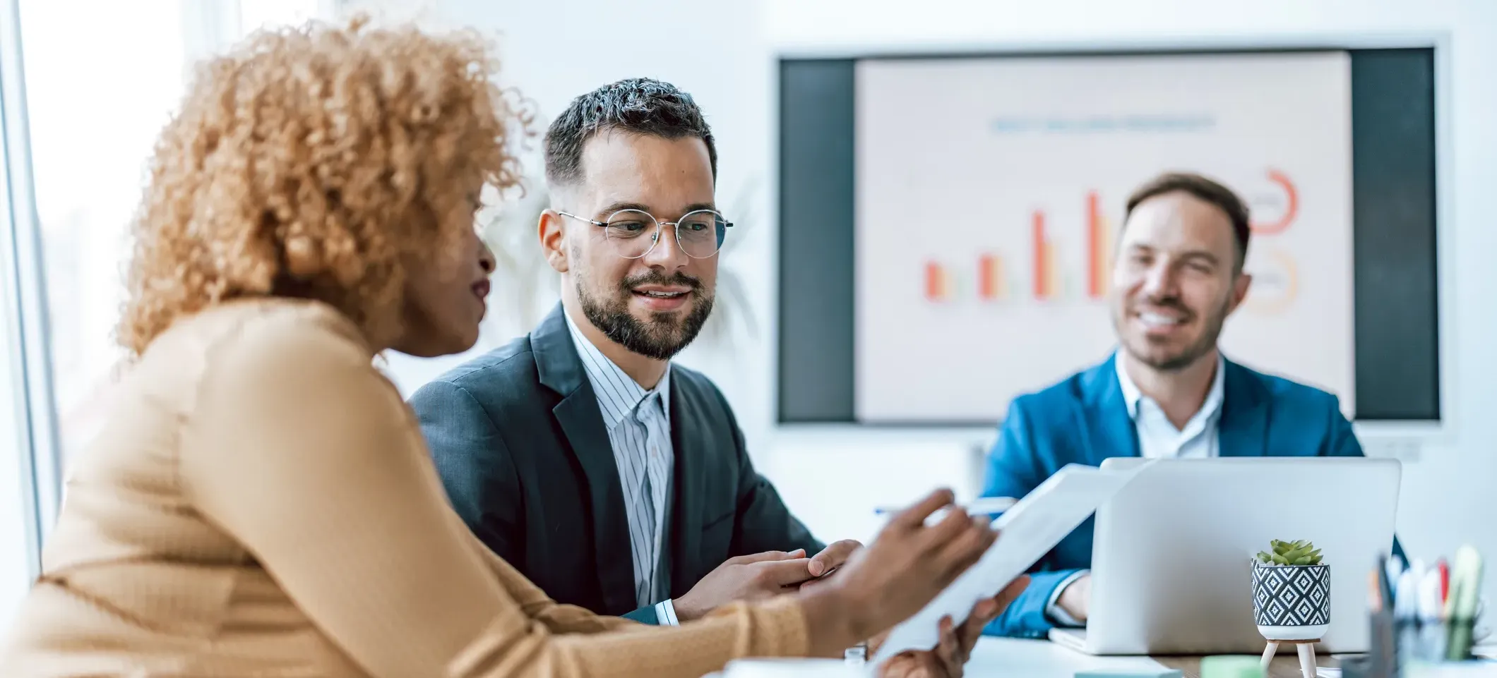 [Featured image] Three business professionals are sitting at a table looking at a document and discussing KPI tracking.
