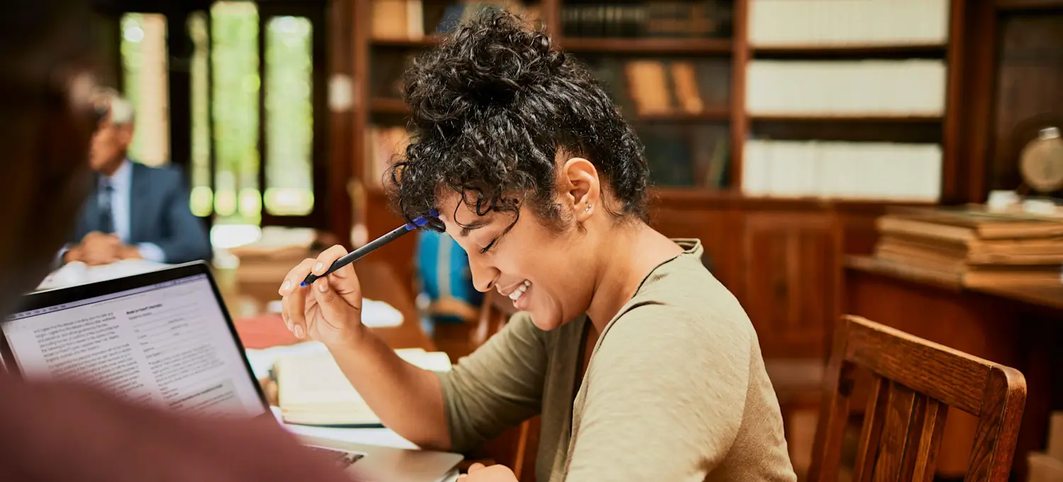 [Featured Image] A person in a library is holding a pen to their temple and looking at their laptop as they study for their PhD.