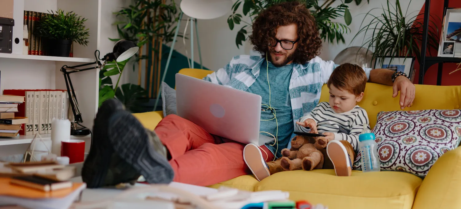 [Featured Image]: A nontraditional student sits on a yellow sofa with a young toddler next to them. They are working on a college course on their laptop computer.