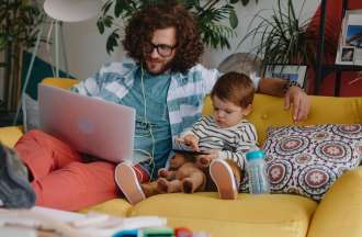 A man sits on a yellow sofa wit ha young toddler next to him. He's working on a college course on his laptop computer.