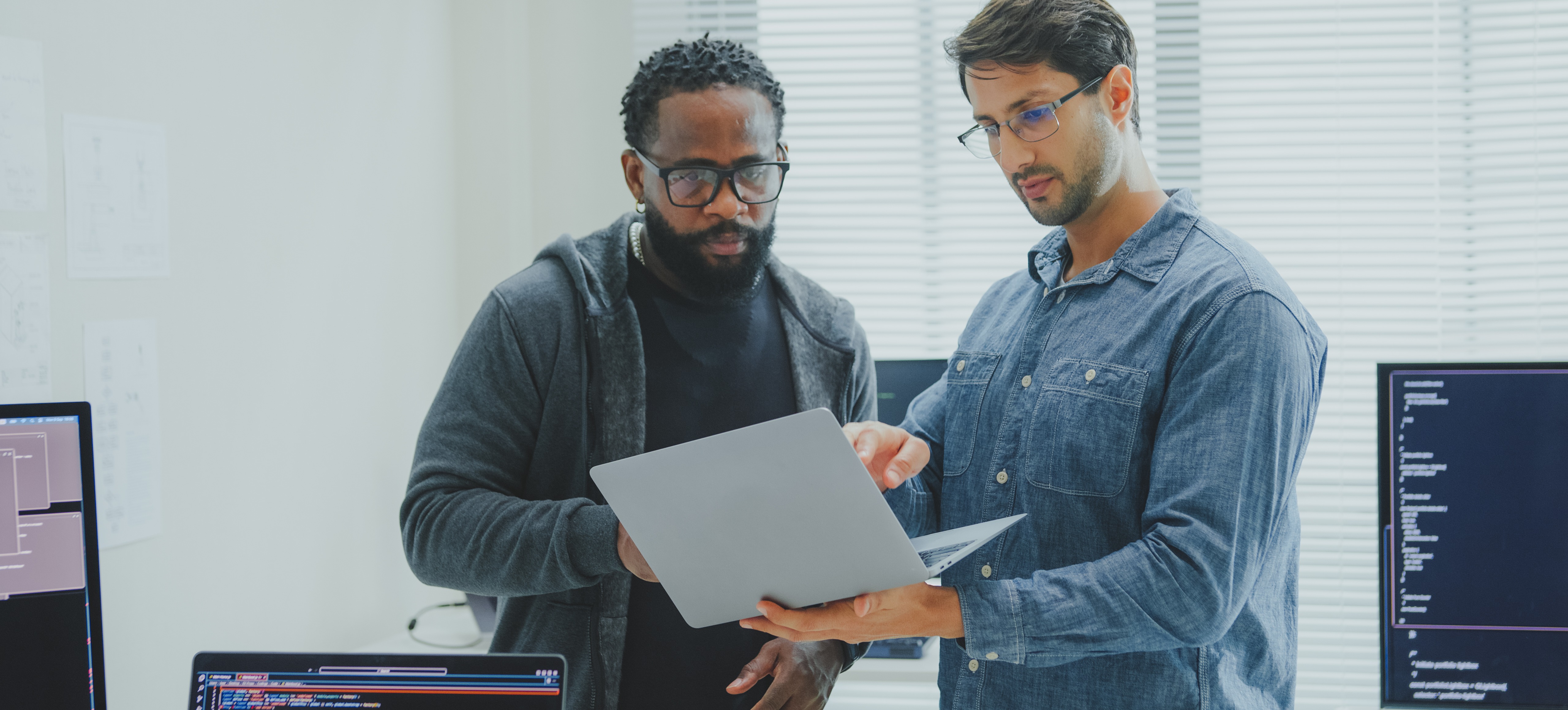 [Featured Image] Two data scientists stand in an office and discuss causal machine learning while looking at a computer screen.
