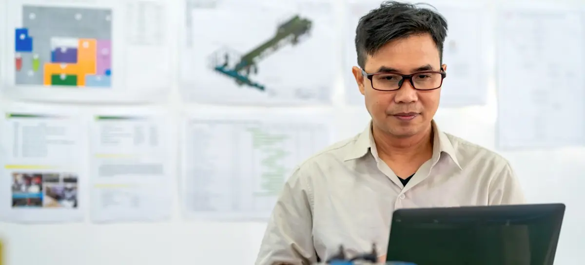 [Featured image] A Master of Engineering student wearing glasses stands in front of a wall with design specifications on it. They are working on their laptop.