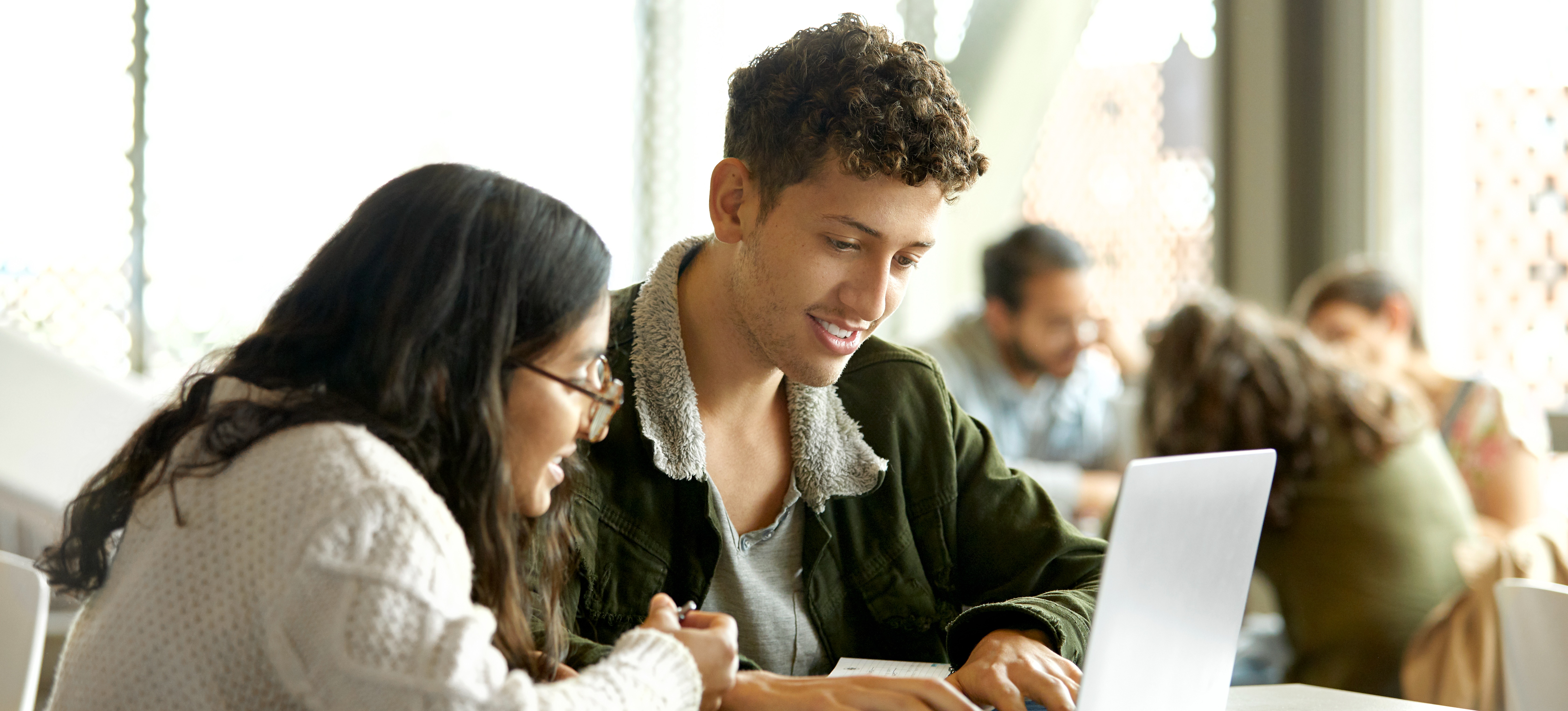 [Feature Image] Two prospective business learners conduct online research, querying, “How long does it take to get a business degree?” on a laptop in a well-lit area with other learners in the background.
